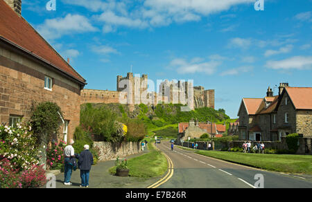 Ansicht des riesigen und historischen Bamburgh Castle auf grasbewachsenen Hügel mit Hütten der Stadt und Menschen im Vordergrund und blauer Himmel in England Stockfoto