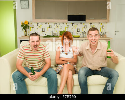zwei Männer sitzen auf der Couch vor dem Fernseher, in Küche, hält Biere und schreien, mit einer verärgert Frau dazwischen Stockfoto