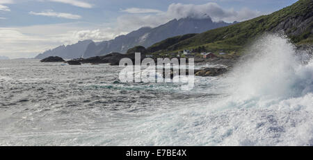 Wellen an der felsigen Küste, Cottages und zerklüftete Berge brechen auf der Rückseite, flakstad, Lofoten, Nordland, Norwegen Stockfoto