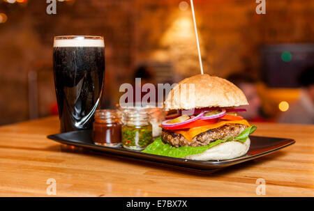 Leckeres Beef-Burger mit Salat, Tomaten, Käse, Zwiebel, Salsa, Ketchup, Pesto mit Dunkelbier auf Holztisch Stockfoto