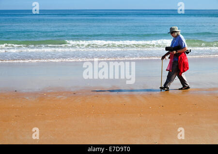 Frau Ormond Strand entlang auf der Suche nach Muscheln. Sie trägt einen Spazierstock, einen Stuhl und einen Beutel Muscheln Stockfoto