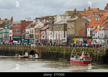 Menge von Menschen flanieren neben Whitby Hafen mit Gebäuden der beliebten englischen Küstenstadt Urlaub gruppierten entlang des Kais Stockfoto