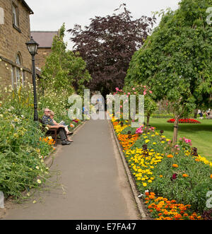 Bunte Anzeige von Frühlingsblumen neben Wiesen und Bäumen mit Menschen entspannen auf Parkbank in Bad Gärten, Bakewell, England Stockfoto