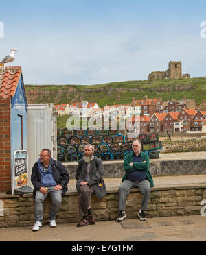 Drei ältere Männer sitzen an Wand neben Hafen an der englischen Küste Stadt Whitby mit Häusern auf Hügel im Hintergrund Stockfoto
