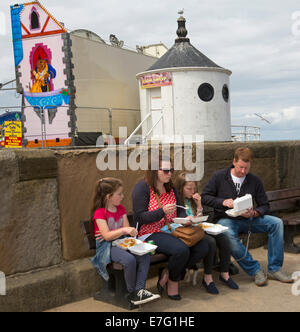 Familie mit zwei Kindern sitzen auf Wand Essen von Fisch und Chips aus emporter Behältern im englischen Küstenort - Whitby Stockfoto