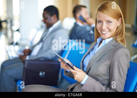 Weibliche Angestellte mit Touchpad Blick in die Kamera mit zwei Männer sitzen auf Hintergrund am Flughafen Stockfoto