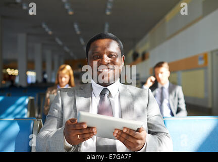 Young African-American Geschäftsmann mit Touchpad sitzen im Flughafen mit zwei Kollegen im Hintergrund Stockfoto