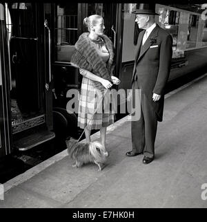 1950 historische Abbildung, die den Stewart oder Maitre d' der Brighton Belle Pullman Waggon auf einem Bahnsteig in London Victoria in einem Gespräch mit einem Smart lady Passagier mit einem Fell um sie herum und mit einem kleinen Hund an der Leine zu werfen. Pullman Züge begannen 1874 und wurden mainline Luxus Railway Services thar wth erstklassige Trainer und eine Stewart Service betrieben. Stockfoto