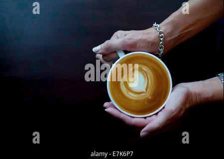 Reife Hände einer Frau mit einem Kaffee Café Latte Cappuccino mit Herz-Design im Schaum Stockfoto