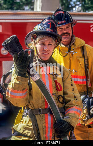 Tragen ihre Feuerwehrhelm und Feuer Mantel, eine San Diego, CA, hält Frau Feuerwehrmann einen Wasserschlauch aus einem Feuerwehrauto. Hinweis männlicher Kollege. Stockfoto