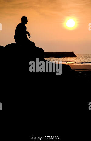 Die Silhouette eines Mannes sitzt auf einem Felsen Sennen Cove wie die Sonne setzt. Stockfoto