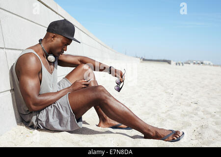 Junger Kerl eine SMS am Strand. Afrikanischer Mann sitzt am Strand mit Handy. Muskulöse männliche Model im Freien. Stockfoto