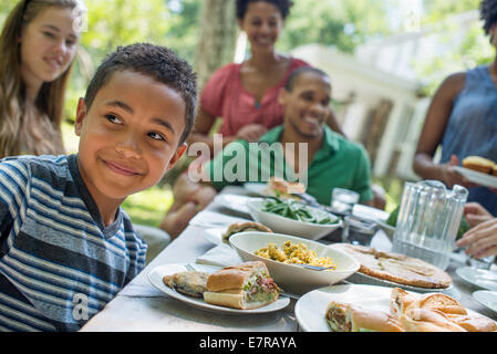 Ein Familientreffen, Männer, Frauen und Kinder an einem Tisch in einem Garten im Sommer. Ein Junge, lächelnd in den Vordergrund. Stockfoto