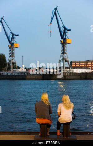 Zwei Frauen sitzen am Hafen an der Elbe mit Kranen im Hintergrund in der Abenddämmerung. Stockfoto