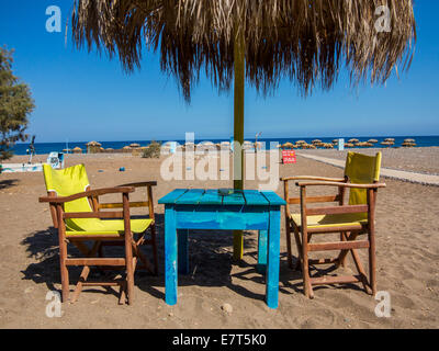 Zwei gelbe, eine blaue Tisch, Stühlen und einem Palm-Unmbrella an einem Sandstrand in der Sonne mit einem strahlend blauen Meer in der Ferne Stockfoto