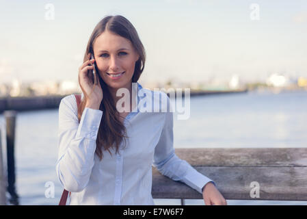 Attraktive Frau, die einen Anruf auf ihrem Handy ansehen im Freien vor dem Fluss Hintergrund lächelt in die Kamera Stockfoto