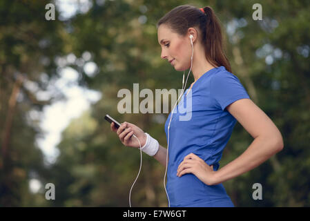 Attraktive glücklich Frau stehend hören Sie Musik auf einem Feldweg durch einen Wald mit ihrem MP3-Player und Ohrstöpsel lächelnd Stockfoto