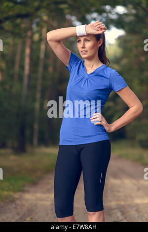 Sportlerin, Schweiß von der Stirn auf ihrem Armband abwischen, da sie während ihrer Übungen auf einem Waldweg macht eine Pause Stockfoto