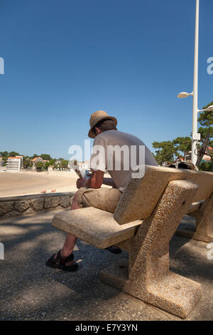 Ein Mann sitzt auf schattigen Bank mit Blick auf Strand, Buch zu lesen. Stockfoto