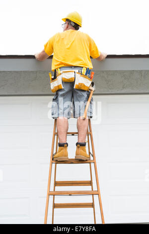 Ein Unternehmer mit einem Gürtel voller Werkzeuge geht auf eine Leiter auf das Dach von einem Heim zu inspizieren. Stockfoto