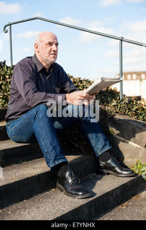 Glatze Mann mittleren Alters in Jeans sitzen im Freien in der Sonne auf einer Treppe eine Zeitung lesen Stockfoto