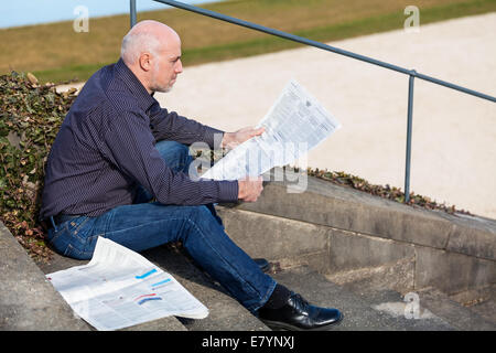 Glatze Mann mittleren Alters in Jeans sitzen im Freien in der Sonne auf einer Treppe eine Zeitung lesen Stockfoto