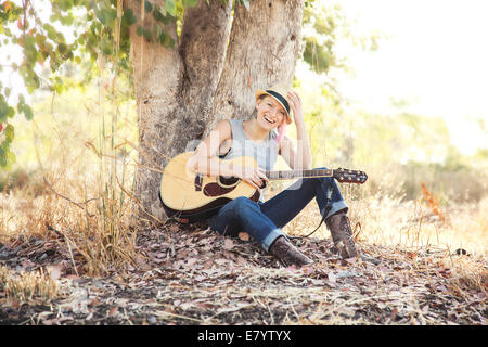 Frau mit Akustikgitarre sitzen unter Baum Stockfoto