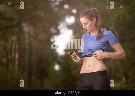 Junge sportliche Frau trainieren im Wald stehen, die ihr Smartphone zu betrachten, wie sie eine mobile Anwendung elektronischer nutzt Stockfoto