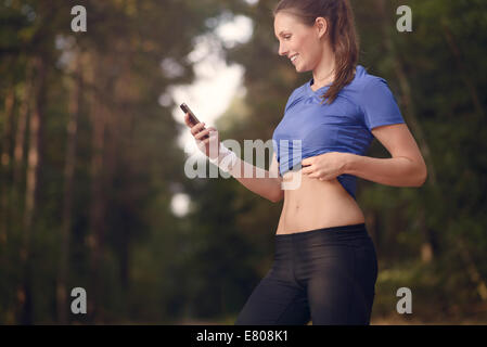 Junge sportliche Frau trainieren im Wald stehen, die ihr Smartphone zu betrachten, wie sie eine mobile Anwendung elektronischer nutzt Stockfoto