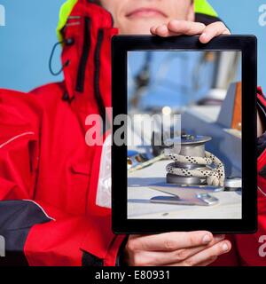 Mann in Windjacke hält Ipad mit Foto von Yacht Segelboot. Seemann, zeigt der Bildschirm Tablet Touchpad träumen Yachting Segel Stockfoto