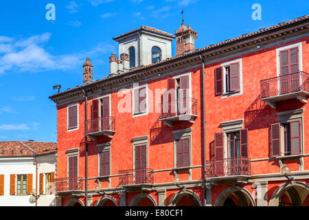 Fassade des roten, zweistöckiges Gebäude mit Balkonen und Fensterläden aus Holz unter blauem Himmel in Alba, Italien. Stockfoto