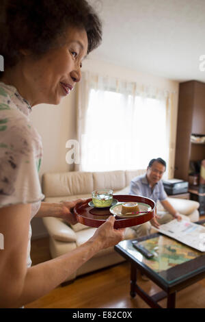 Eine Frau, ein Tablett mit Essen, einem Mann sitzt auf einem Sofa dienen. Stockfoto