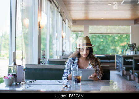 Eine junge Frau sitzt an einem Tisch in einem Diner, trägt einen Strohhut mit einem digitalen Tablet. Stockfoto