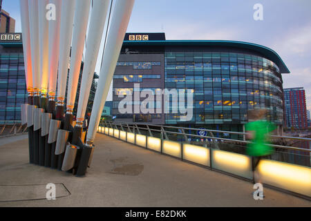 BBC's Flaggschiff-Fernsehzentrale im Norden in MediaCity, Media City, Salford Quays mit einem Jogger, der die MediaCityUK Link Fußgängerbrücke überquert, die im Morgengrauen beleuchtet wird. Stockfoto