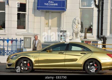 Ein Alter Mann hört und schaut auf eine Gold lackiert Mercedes Benz Sport-Limousine auf Eastbourne Strandpromenade geparkt Stockfoto