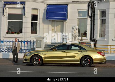 Ein Alter Mann hört und schaut auf eine Gold lackiert Mercedes Benz Sport-Limousine auf Eastbourne Strandpromenade geparkt Stockfoto