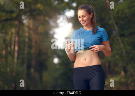 Junge sportliche Frau trainieren im Wald stehen, die ihr Smartphone zu betrachten, wie sie eine mobile Anwendung elektronischer nutzt Stockfoto