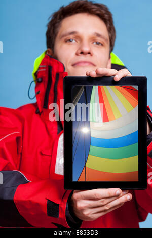 Mann in Windjacke hält Ipad mit Foto von bunten Segel Boot. Zeigt der Bildschirm Tablet Touchpad träumen über Yacht Segler Stockfoto
