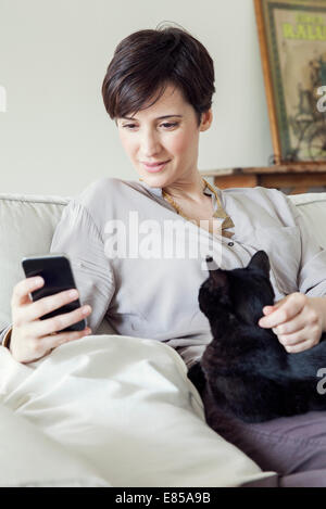 Frau sitzt auf dem Sofa mit Katze auf dem Schoß, mit smartphone Stockfoto