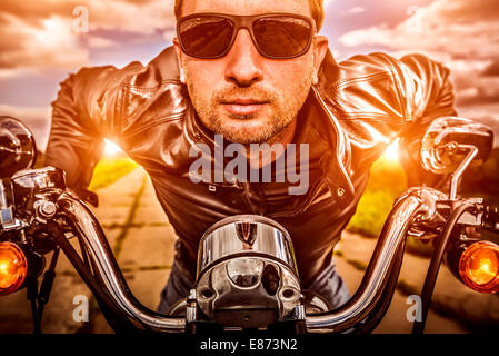 Biker-Mann trägt eine Lederjacke und eine Sonnenbrille sitzt auf seinem Motorrad, Blick auf den Sonnenuntergang. Filter in der Postproduktion angewendet. Stockfoto