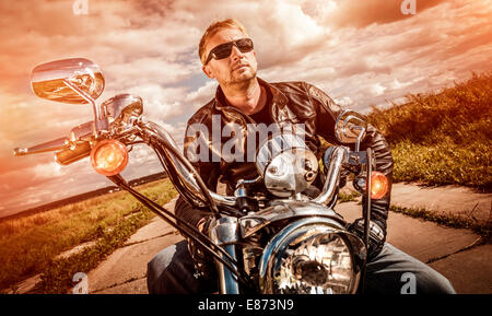 Biker-Mann trägt eine Lederjacke und eine Sonnenbrille sitzt auf seinem Motorrad, Blick auf den Sonnenuntergang. Stockfoto