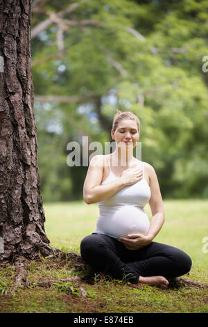 Porträt von weißen schöne schwangere Frau Yoga trainieren im Park in der Nähe von Baum, Herz und Bauch zu berühren Stockfoto
