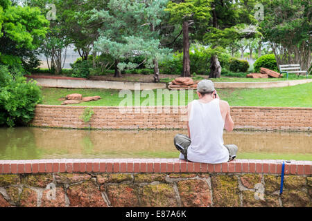 Ein junger kaukasischen Mann sprechen auf einem Handy sitzt auf einer Mauer in Will Rogers Park, Oklahoma City, Oklahoma, USA Stockfoto
