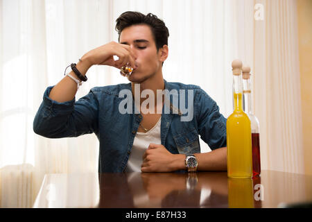 Junger Mann sitzen trinken allein an einem Tisch mit zwei Flaschen Schnaps neben ihm schlürfen von Shot-Glas, seinen Kummer zu ertränken Stockfoto