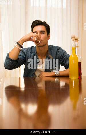 Junger Mann sitzen trinken allein an einem Tisch mit zwei Flaschen Schnaps neben ihm schlürfen von Shot-Glas seine ertrinken Stockfoto