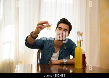 Junger Mann sitzen trinken allein an einem Tisch macht einen Toast mit zwei Flaschen Schnaps neben ihm schlürfen von Shot-Glas Stockfoto