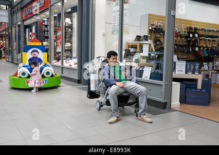 Gelangweilt asiatischen Vater sitzt in seinem Töchter Buggy, spielt mit seinem Smartphone & nicht gerade sein Kind beim Einkaufen Stockfoto