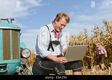 Bauer mit Laptop auf Feld mit Traktor, seinem Sohn spielt im Hintergrund, Bayern, Deutschland Stockfoto