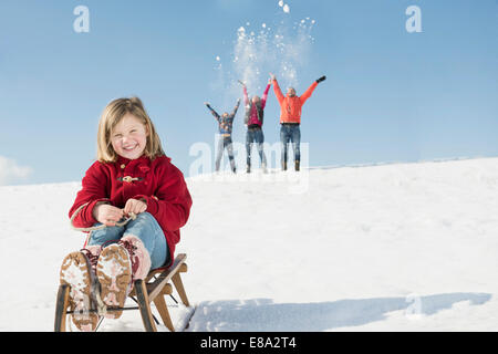 Porträt von Mädchen sitzen auf Schlitten, Familie, spielen im Hintergrund, Bayern, Deutschland Stockfoto