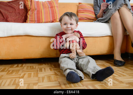 Junge sitzt mit Teddybär im Arm auf Boden, Lächeln Stockfoto